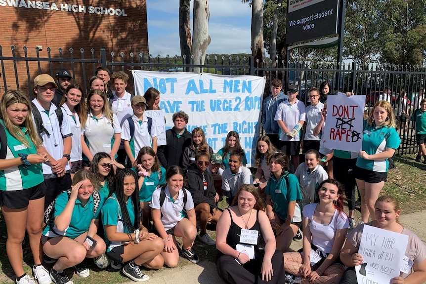 A group of students gather at the school gate with a sign that says: "Not all men have the urge to rape".