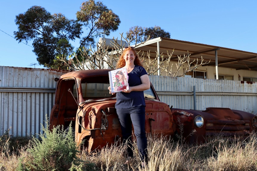 female writer dressed in blue stands in front of two old rusty car showing her manuscript 