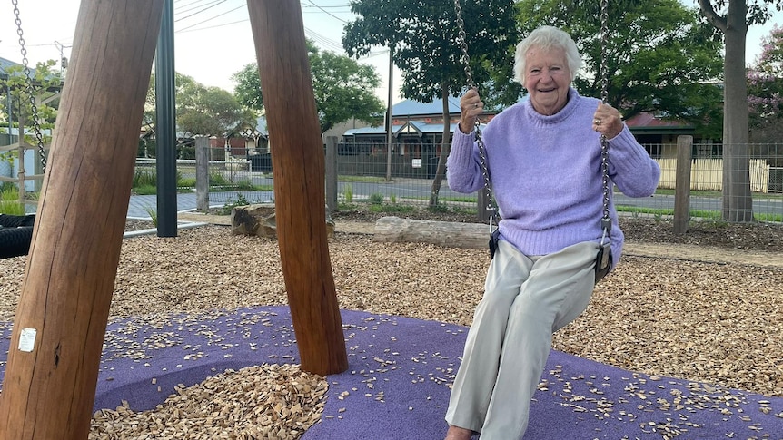 elderly woman sitting on playground swing wearing a purple knitted jumper