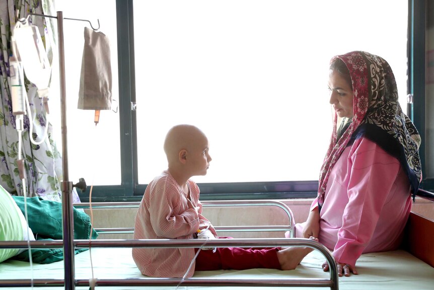 A 5-year-old boy with no hair suffering from eye cancer speaks with his mother on a hospital bed.