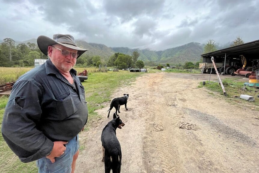 A man in work clothes and a hat on his rural property with two dark coloured dogs.