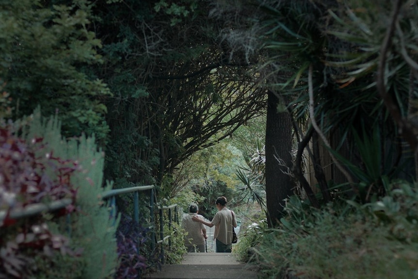 A boardwalk through a lush garden with foilage providing an arch overhead. In the distance, two women, one with a cane.