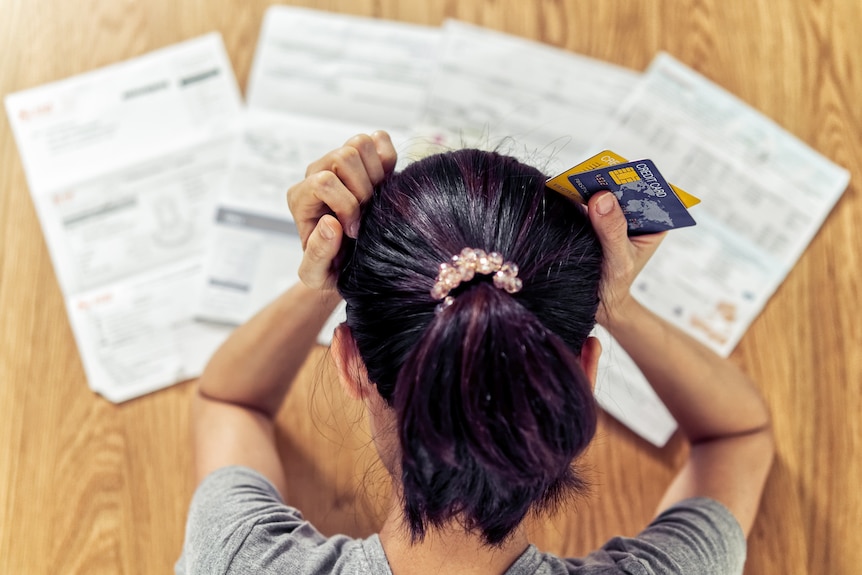 A woman sits at a table covered in bills with her head in her hands, holding two credit cards 
