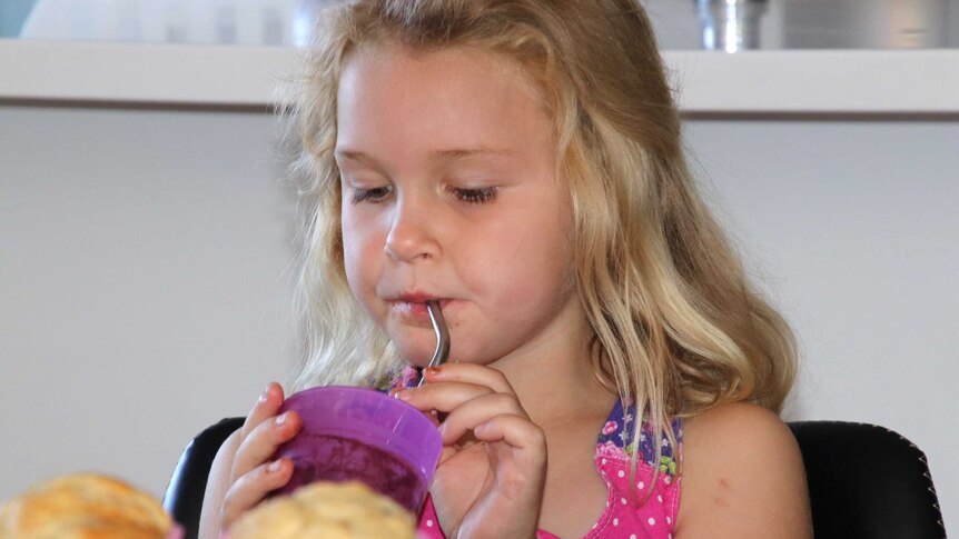A girl with blonde hair sits indoors drinking from a purple cup using a stainless steel straw.
