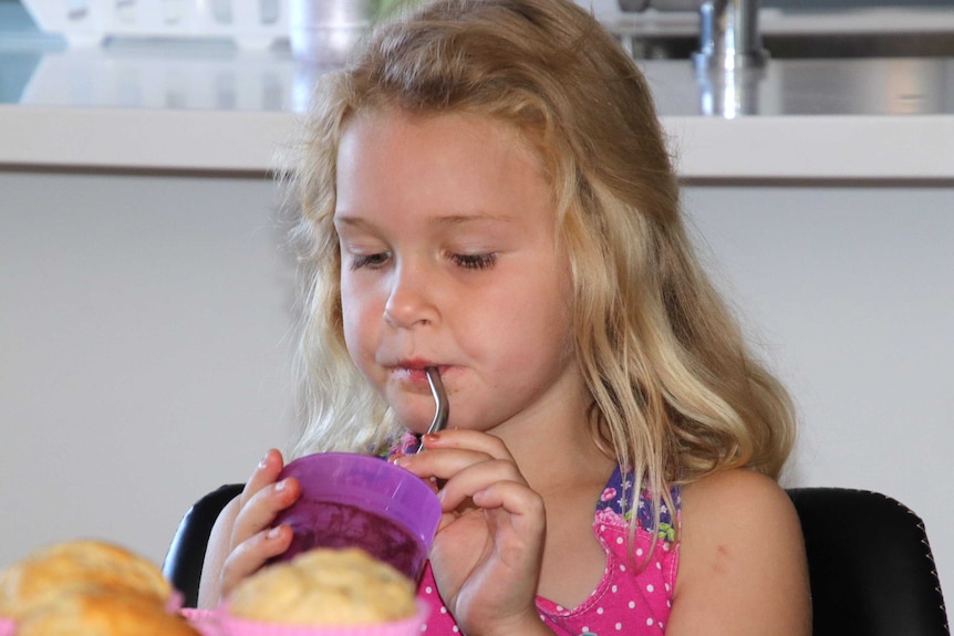 A girl with blonde hair sits indoors drinking from a purple cup using a stainless steel straw.