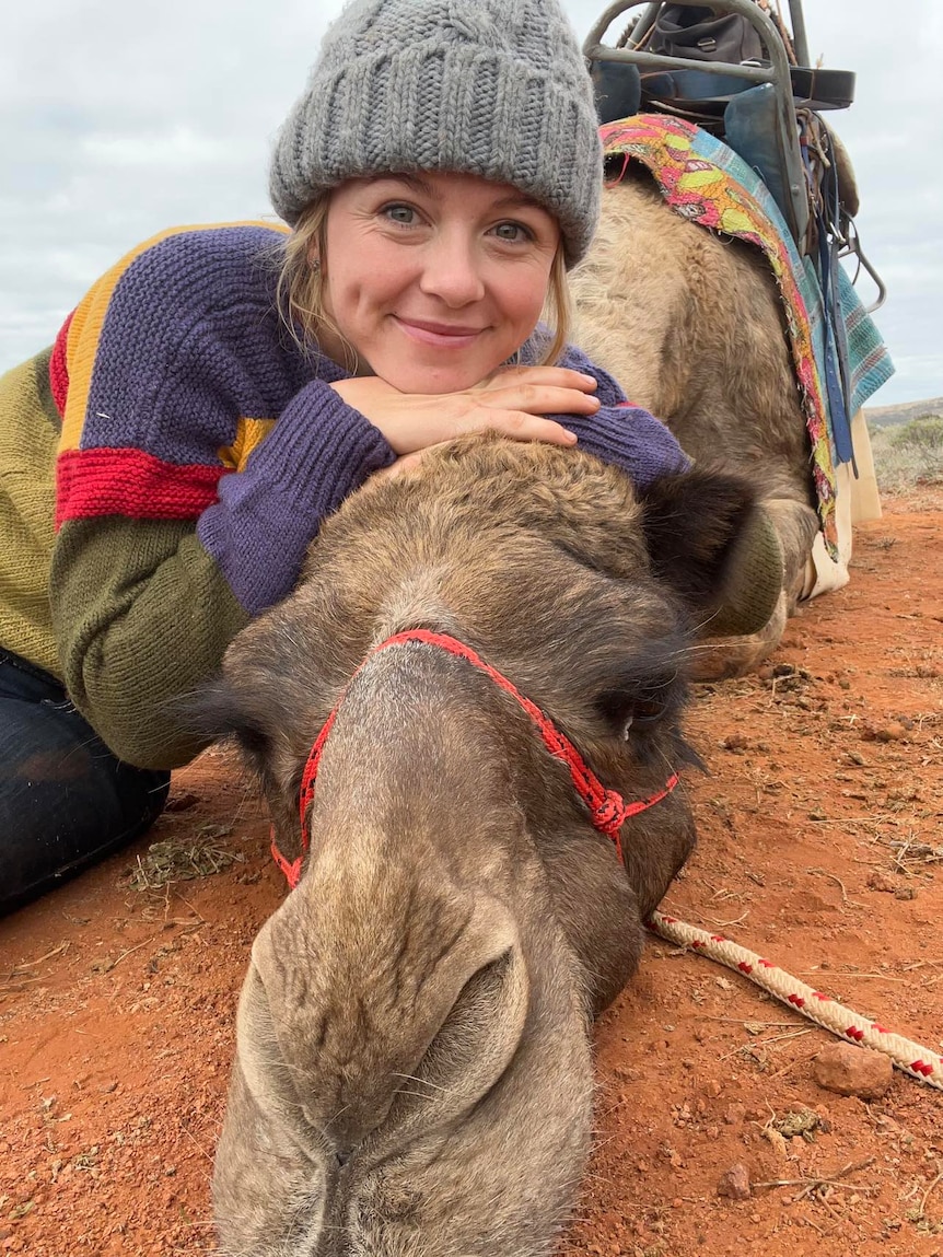 a woman in a beanie resting her head on a camel