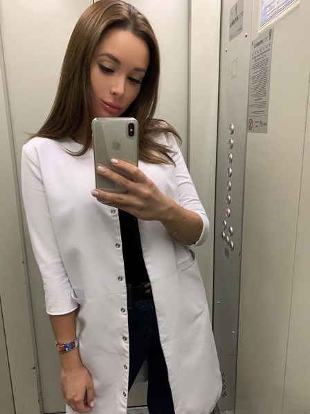 A woman takes a photo with her phone in a lift. She is wearing a white lab coat.