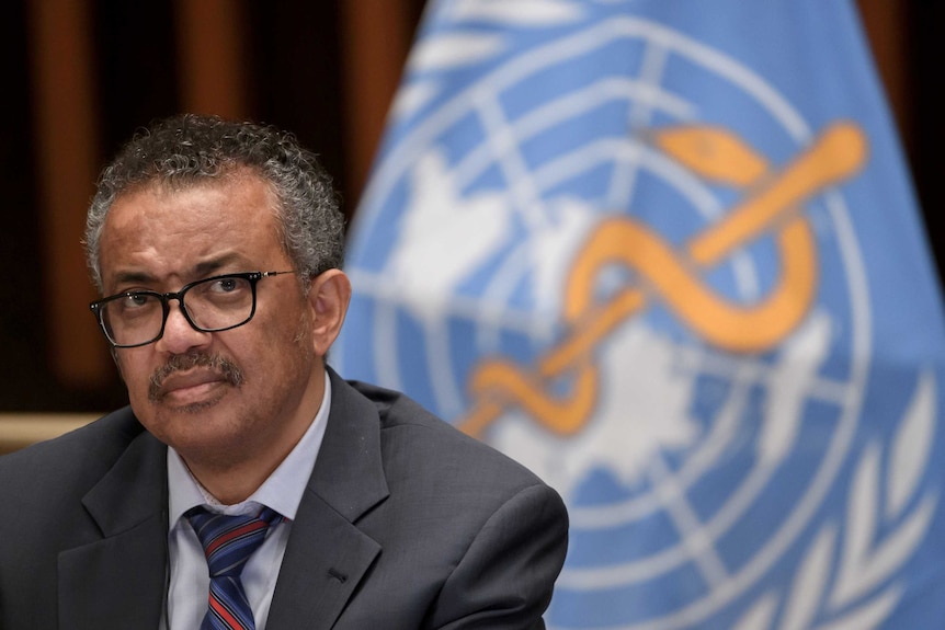 a man with glasses and a moustache in a suit sits in front of the world health organization logo