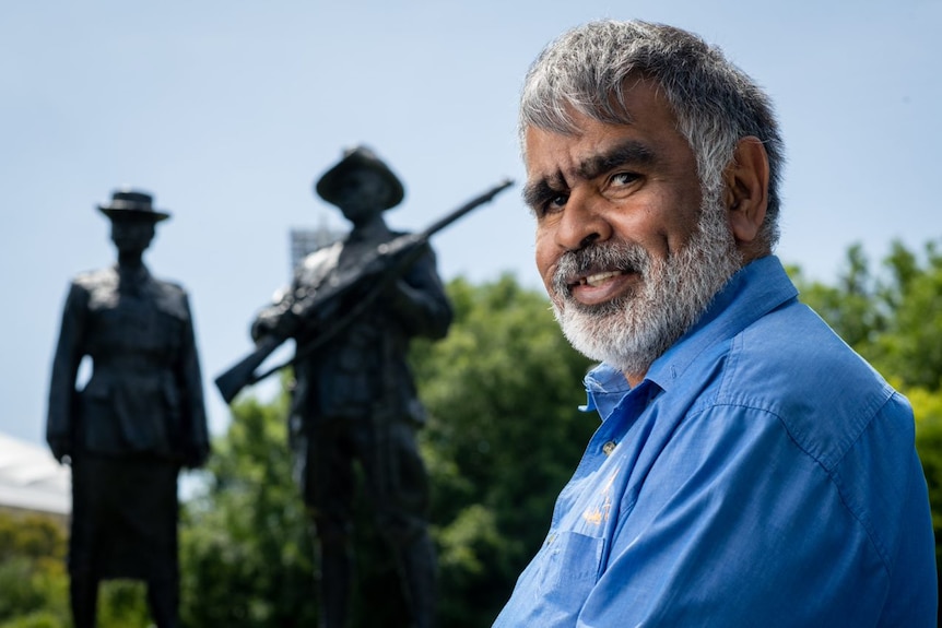An Aboriginal man stands side on smiling at the camera with colonial statues out of focus in the background