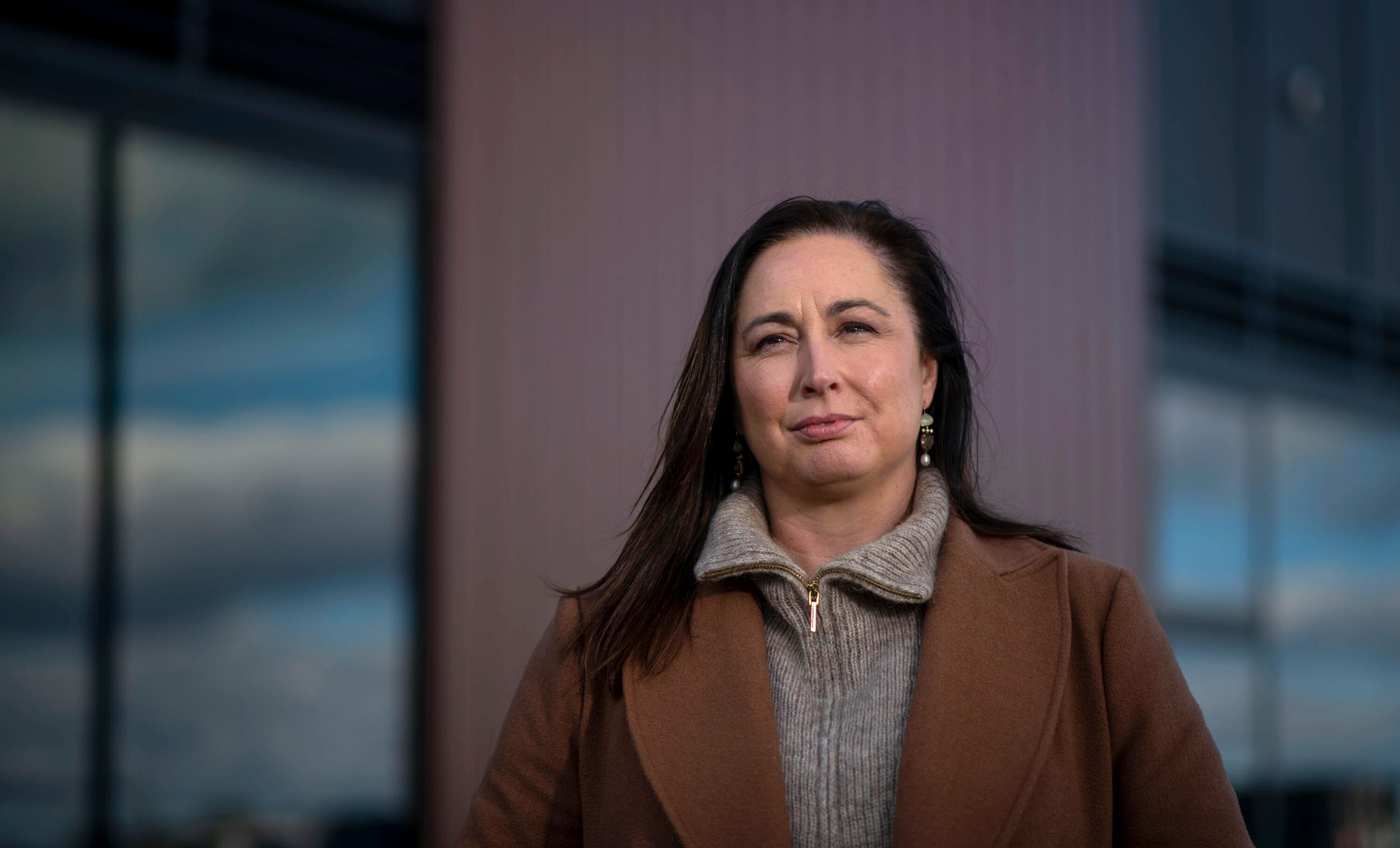 A woman with dark hair and a brown jacket outside metallic office buildings under a blue sky.