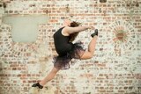 A ballerina dancer wearing a black suit jumps in the air with a textured brick wall behind her