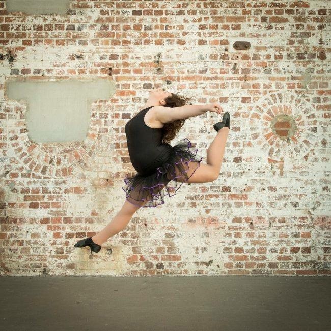 A ballerina dancer wearing a black suit jumps in the air with a textured brick wall behind her