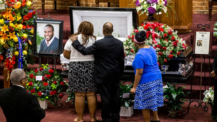 A man wraps his arm around the shoulder of a woman as they look into an open casket surrounded by flowers and photographs
