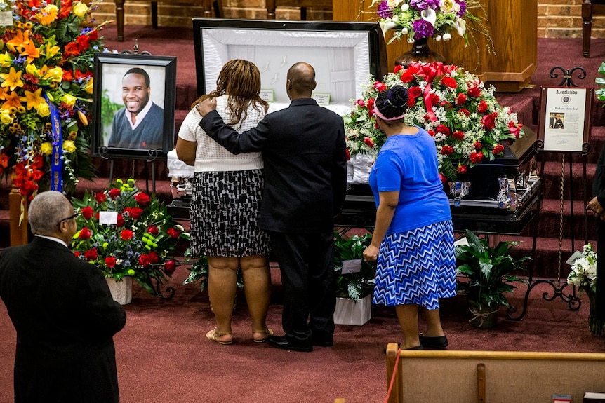 A man wraps his arm around the shoulder of a woman as they look into an open casket surrounded by flowers and photographs
