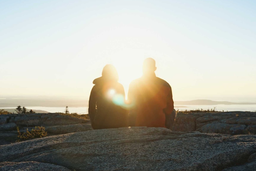 Two people sit on a rock against a blue sky with their backs to the camera while the sun's rays shines between them.