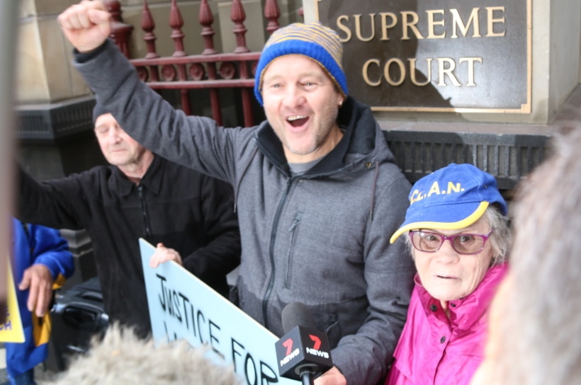 A man in a grey hoodie and a yellow and blue beanie cheers with his fist in the air outside the Supreme Court