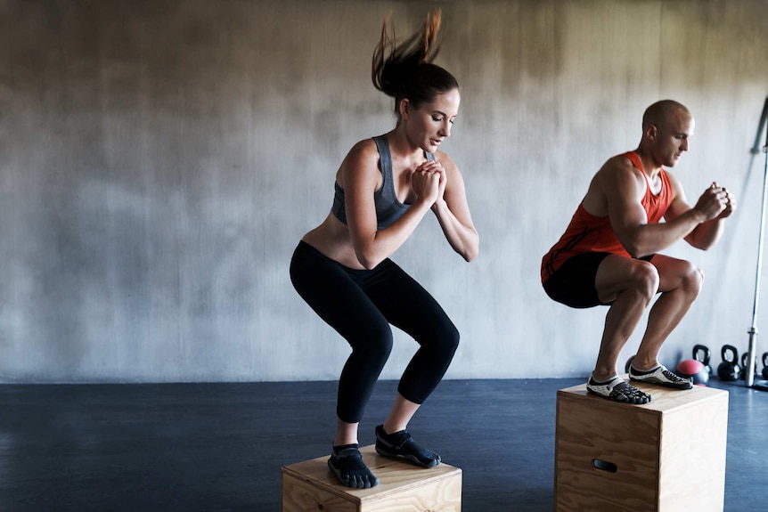 A man and a woman wearing fitness gear jump onto boxes