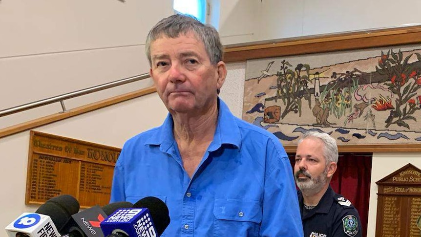 A man wearing a blue shirt and jeans stands in front of microphones on stands