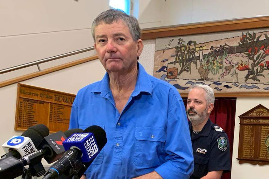 A man wearing a blue shirt and jeans stands in front of microphones on stands