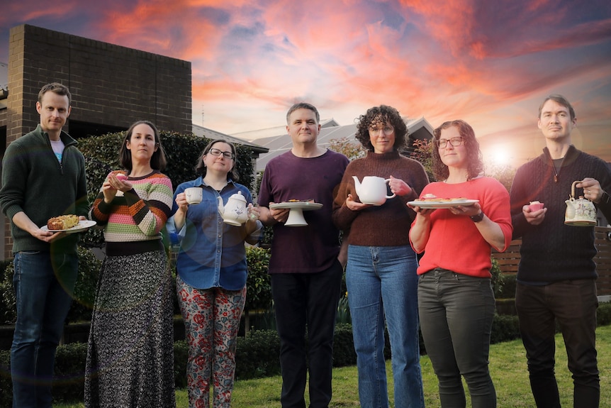 Seven people stand on a lawn in front of a sunset, holding teapots, tea cups and cakes.