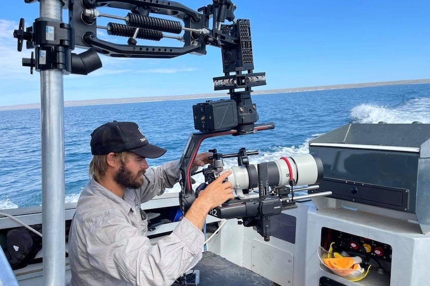 Man operating a camera on a boat. It's a clear, blue day and he is surrounding by gear and rigging.