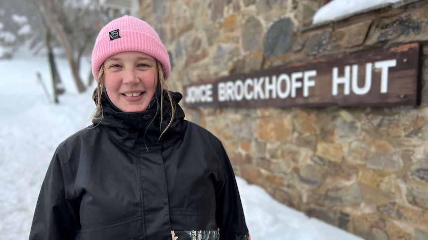 A young, fair-haired woman wearing a pink beanie and a heavy jacket stands in the snow at Joyce Brockhoff Hut