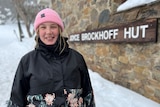 A young, fair-haired woman wearing a pink beanie and a heavy jacket stands in the snow at Joyce Brockhoff Hut