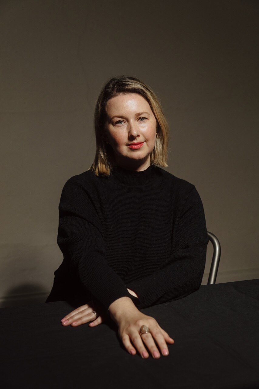 A middle-aged white woman with blonde hair wearing black, sitting with her arms crossed in front of her