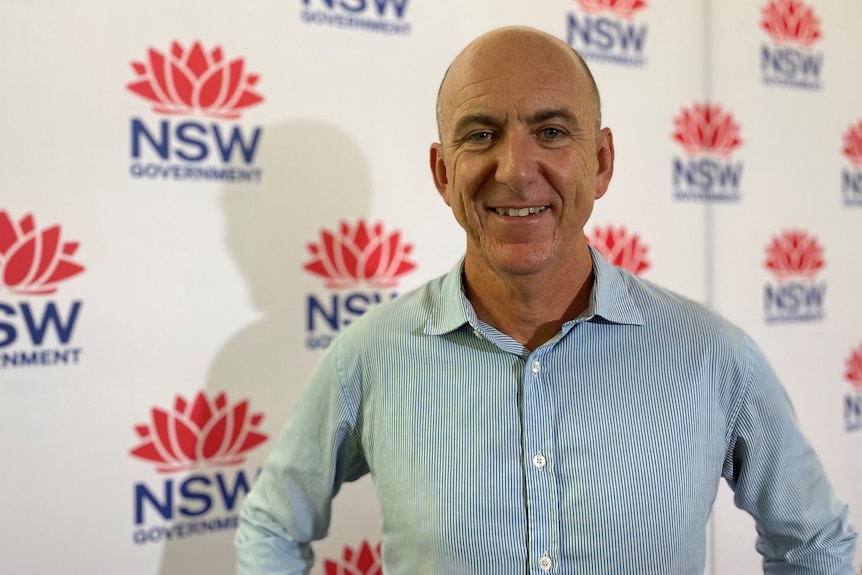 A bald man in a collared shirt stands in front of a backdrop featuring NSW Government branding.