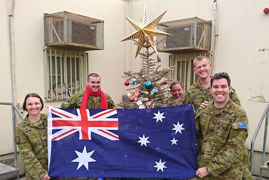 Australian Defence Force members hold up an Australian flag in front of a Christmas tree.