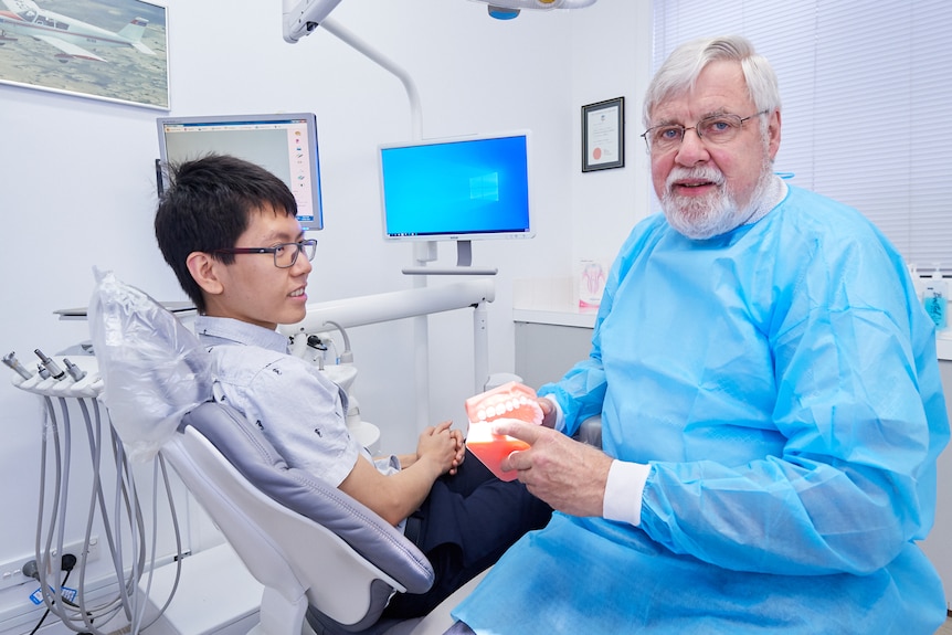 A man wearing a blue medical gown sits next to a young man in a dental chair. He's holding a model of the mouth.