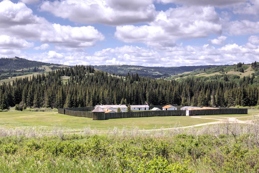 The Northwest Mounted Police fort in Saskatchewan, Canada is shown fenced in a green field, surrounded by rolling hills.