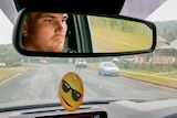 A man's face reflects in the rear-view mirror of the car he is driving.
