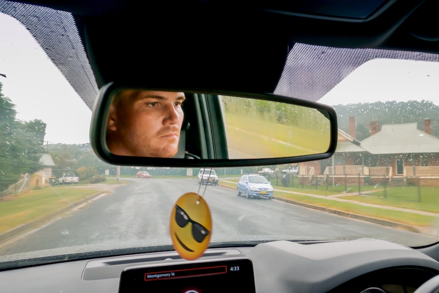 A man's face reflects in the rear-view mirror of the car he is driving.