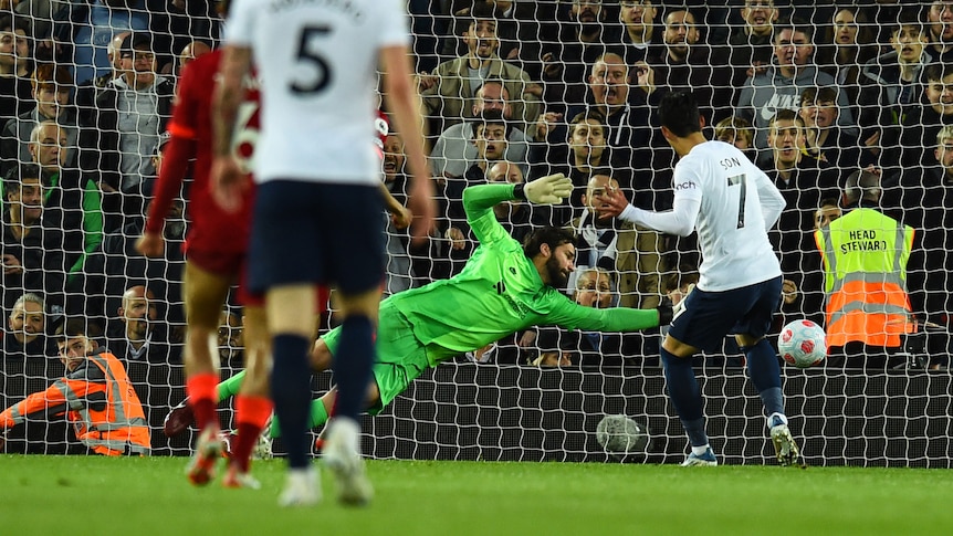 A Premier League striker watches the ball go wide of a diving goalkeeper into the net as fans stare back.