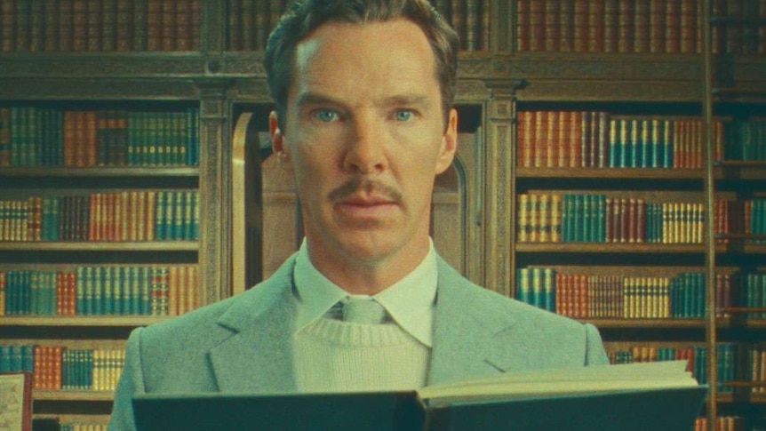 A sepia-toned image of Benedict Cumberbatch in a blue suit reading from a book in front of a wall of bookshelves.