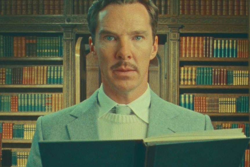 A sepia-toned image of Benedict Cumberbatch in a blue suit reading from a book in front of a wall of bookshelves.