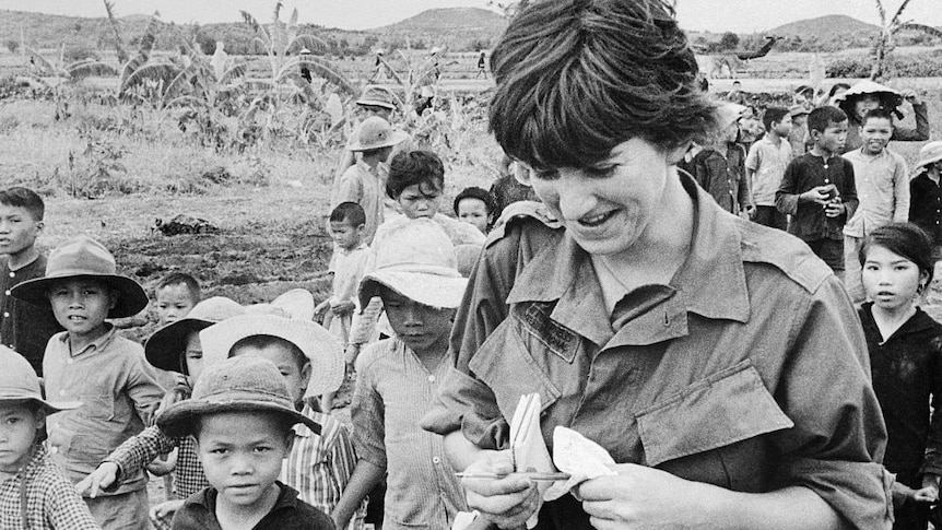B&W of Kate Webb smiling, pen and notepad in hand surrounded by Vietnamese children
