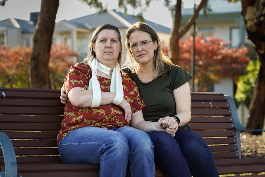 two women holding hands, one with an arm sling, sitting on a park bench