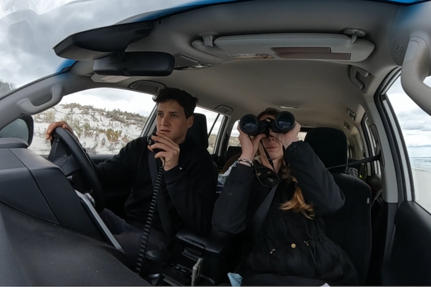 The pair are photographed with a GoPro in the front of a car. Katy has binoculars pressed to her face, Max speaks into a UHF 