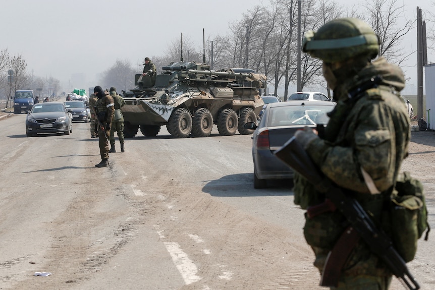 Men in military camo stand guard of a road. Two cars approach.