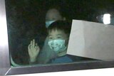 A close-up shot of a white bus and a small boy wearing a face mask waving through the window.