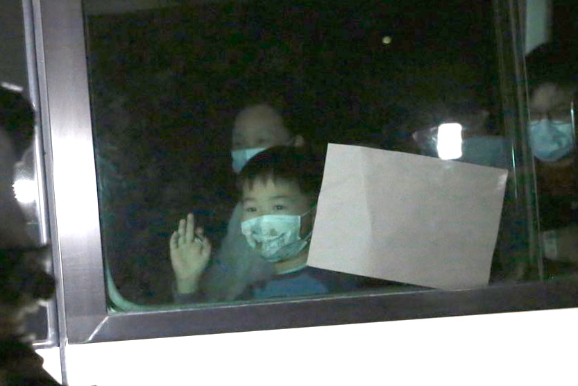 A close-up shot of a white bus and a small boy wearing a face mask waving through the window.