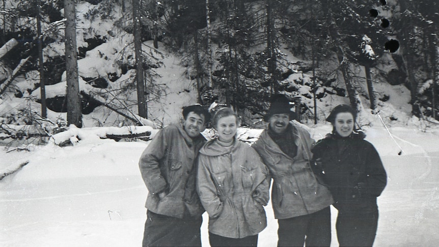A black and white photo of four young people standing together smiling in a snowy forest 