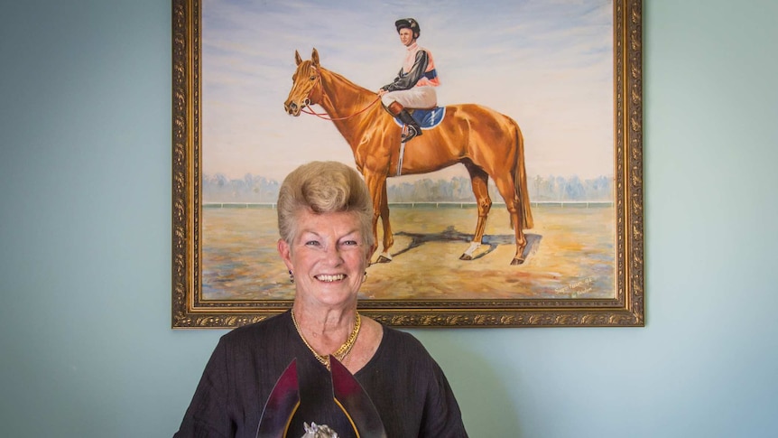 Woman holding a cup underneath a painting of her as a jockey.