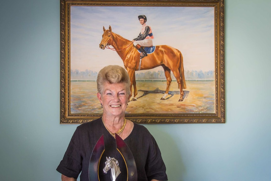 Woman holding a cup underneath a painting of her as a jockey.