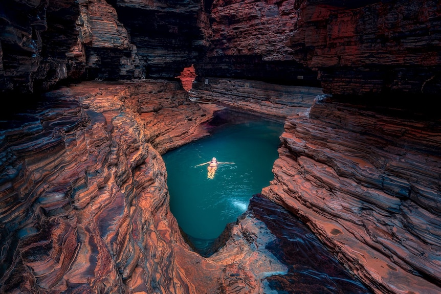 A deep blue pool of water surrounded by rocks, with a woman swimming on her bck.