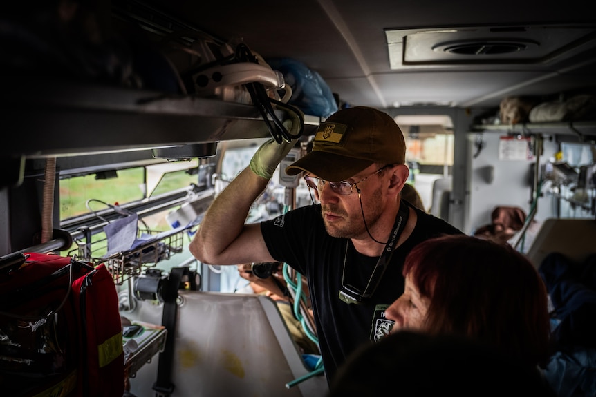 A middle-aged man wearing glasses, cap and medical gloves grabs onto a handle inside a bus kitted out with hospital beds