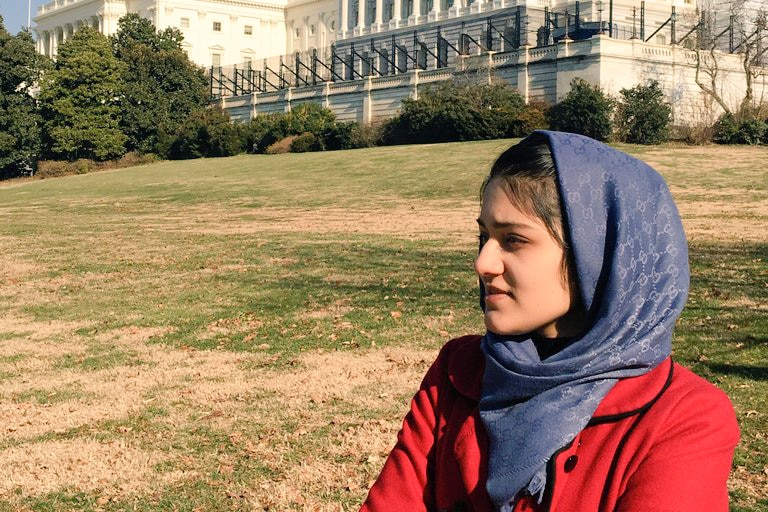 A woman wearing a black and white hijab and red jacket looks away infront of a white building.