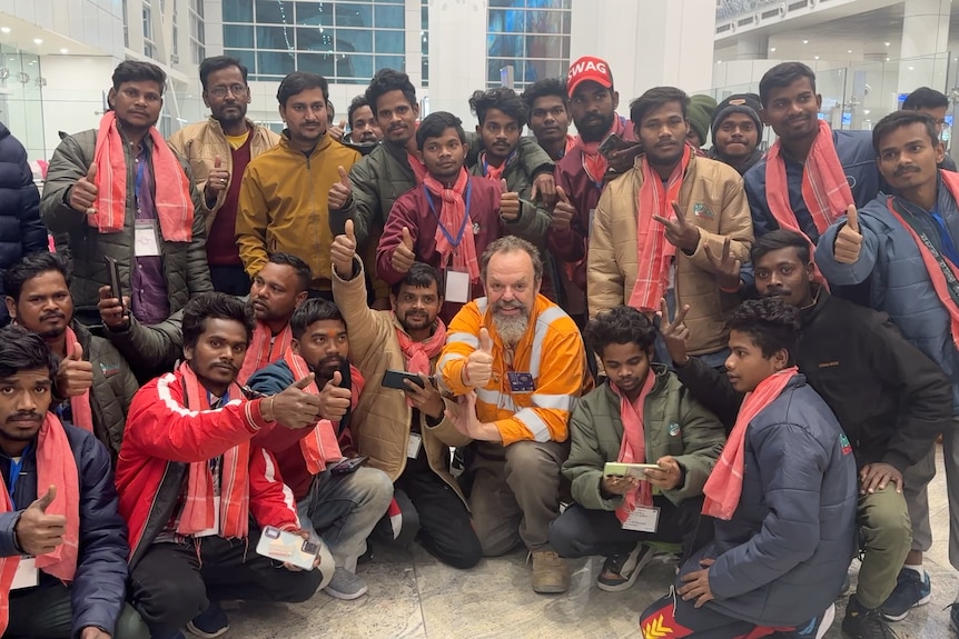 A bearded man in an orange high-vis shirt gives a thumbs up surrounded by 17 Indian men in matching uniforms 